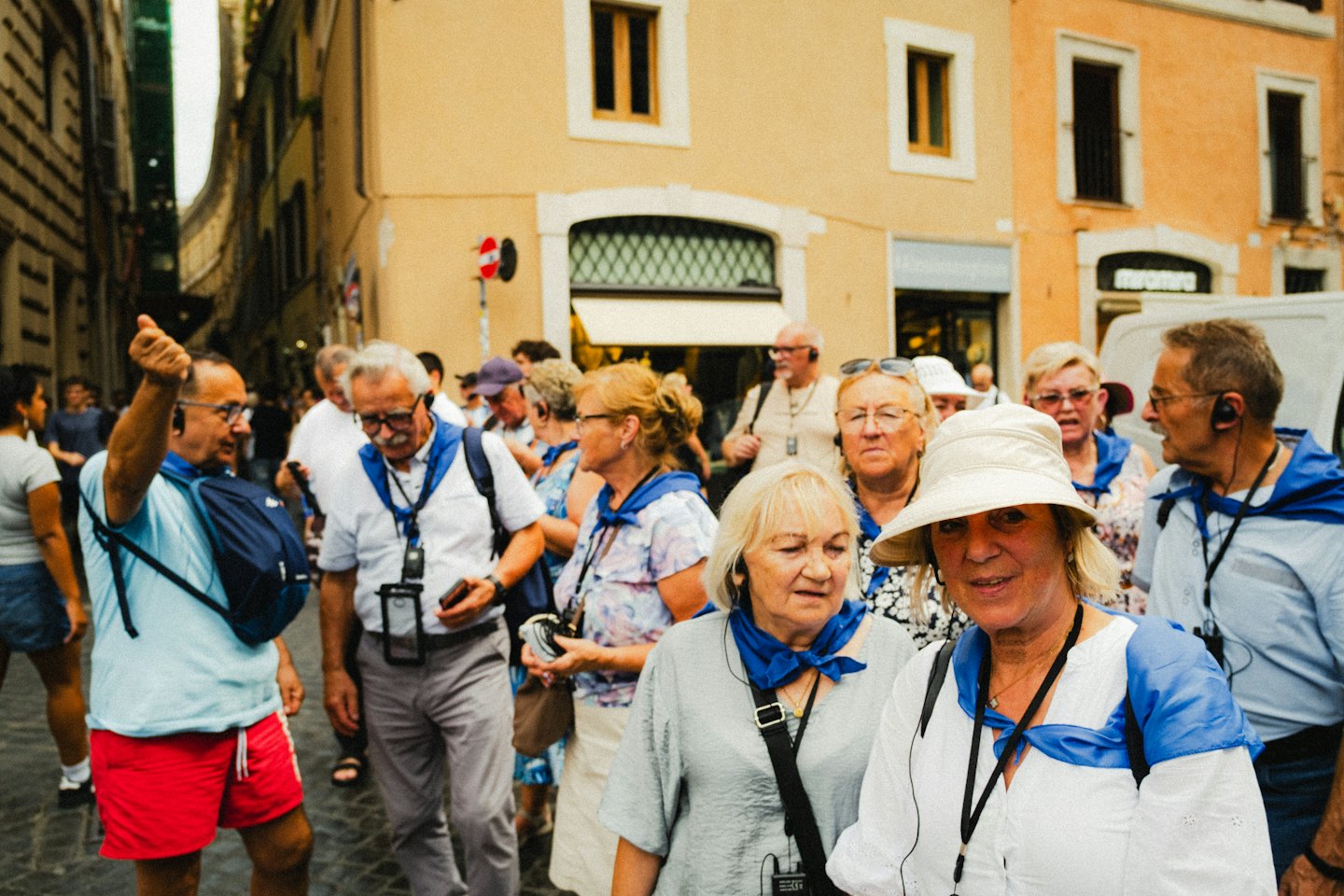 Tourists in blue scarves explore a sunny, narrow street.